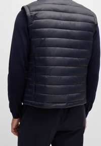 Laivastonsininen puhallettava liivi, jossa on vaakasuora saumaus, kevyt materiaali, hihaton malli ja sileä, kiiltävä pinta.