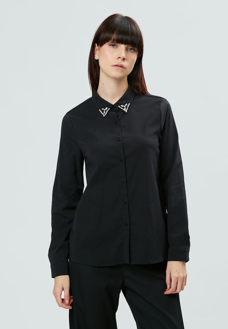 Camisa negra de manga larga con botones en el frente y un collar adornado con detalles decorativos. Fabricada en tejido suave con un corte entallado.