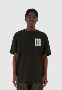 Svart bomull T-skjorte med korte ermer og en stor, stilisert sølvfarget "m" logo på venstre brystområde. Løs passform og rund hals.
