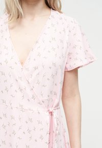 Γυναίκα που φορά ένα ανοιχτό ροζ φόρεμα τύπου wrap με μικρά πράσινα floral σχέδια και κοντά μανίκια με βολάν, δεμένο στη μέση, σε λευκό φόντο.