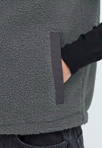 Grau gefütterte Weste mit strukturierten Oberflächen, die eine Seitentasche mit schwarzem Rand aufweist. Lange schwarze Ärmel sind am Handgelenk sichtbar.