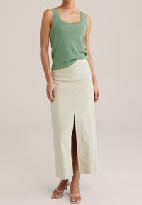 Grünes geripptes Tanktop kombiniert mit einem langen, hellbeigen Rock mit einem Schlitz vorne. Das Model trägt nude-farbene Riemchensandalen.