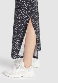 Jambe vêtue d'une jupe noire à motif de petites taches blanches avec une fente latérale, associée à des baskets blanches et grises sur fond clair.