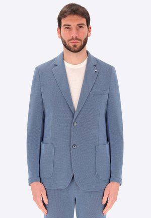 Uomo che indossa una giacca da completo azzurro chiaro testurizzata e pantaloni abbinati sopra una t-shirt bianca con collo rotondo, in piedi su uno sfondo semplice.