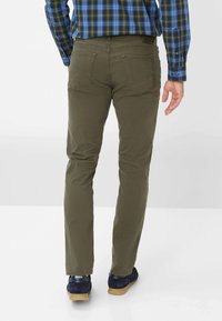 Pantalons slim en vert olive avec des poches arrière, assortis à des chaussures navy. Le tissu a l'air lisse avec un léger éclat.