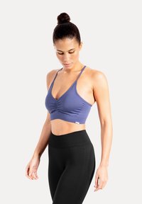 Kvinna med mörkt hår uppsatt i en knut, iklädd en blå sport-bh och svarta, högmidjade leggings, som tittar neråt mot en enfärgad bakgrund.