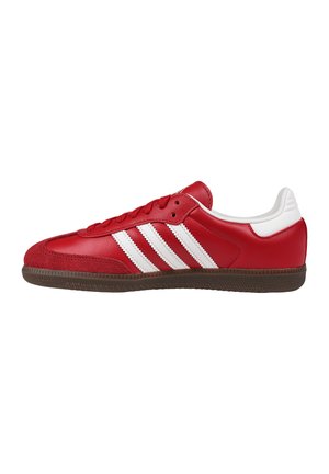 Sneaker bassa rossa con strisce bianche, chiusura frontale con lacci, tab sul tallone bianco e suola di gomma marrone, vista laterale su sfondo bianco.