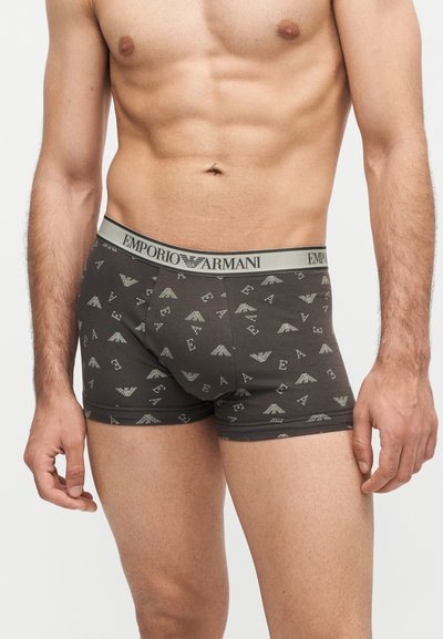 Αρσενικός κορμός που φοράει σκούρα γκρι boxer Emporio Armani με μοτίβο λογότυπου και ζώνη με επωνυμία, στέκεται μπροστά σε λευκό φόντο.