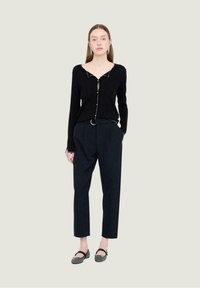 Mujer vestida con un cárdigan negro abotonado, pantalones oscuros de tiro alto con cinturón y zapatos tipo Mary Jane negros, de pie contra un fondo claro.