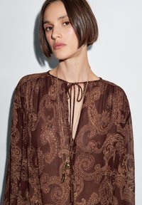 Blusa marrone con motivo paisley, con scollatura a stringere, maniche lunghe e vestibilità ampia, realizzata in tessuto leggero. Indossata con collane sovrapposte.