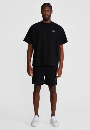 Mężczyzna stojący w czarnej, oversize'owej koszulce i czarnych szortach z białymi trampkami na jednolitym szarym tle.