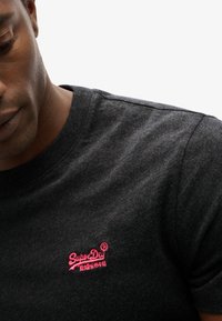 Man in een donkergrijs T-shirt met een klein rood SuperDry-logo geborduurd op de linkerkant van de borst, tegen een witte achtergrond.