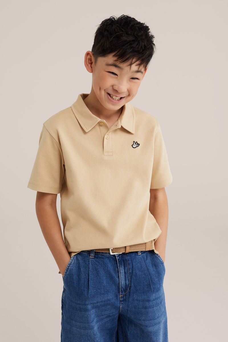 Jeune garçon souriant aux cheveux courts et noirs portant un polo beige et un jean bleu, debout les mains dans les poches devant un fond uni.