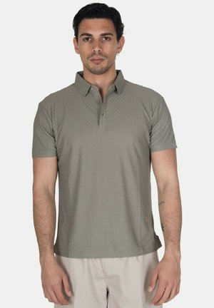 Uomo che indossa una polo verde oliva a maniche corte e pantaloni beige chiaro, in piedi su sfondo bianco uniforme.