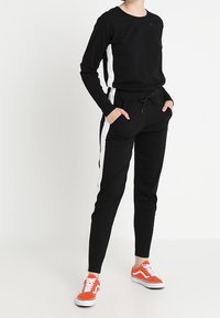 Černý top s dlouhým rukávem a bílými bočními pruhy, kombinovaný s černými joggery s kapsami. Na nohou má oranžové tenisky. Jednoduchý, pohodlný design.
