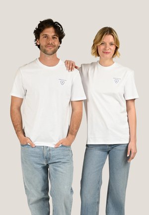 Un homme et une femme se tiennent côte à côte, portant des T-shirts blancs à motifs graphiques et des jeans bleus ; la main de la femme repose sur l’épaule de l’homme.