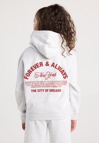 Joven con cabello rizado que lleva un sudadera gris claro y pantalones de chándal, texto en la parte posterior: "Por Siempre y para Siempre Nueva York, La Ciudad de los Sueños," en letras rojas.