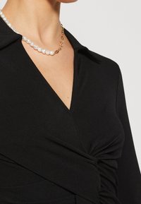 Femme portant une robe portefeuille noire à décolleté en V avec un collier visible composé de perles et d’une chaîne en or autour du cou.