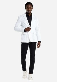 Weißes Struktur-Blazer über einem schwarzen Hemd, kombiniert mit schwarzen Hosen und weißen Sneakers. Der Blazer hat einen Ein-Knopf-Verschluss und Vordertaschen.