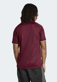 Maroon athletic shirt featuring a geometric pattern, short sleeves, and a gold accent on the collar. Material appears smooth and lightweight.