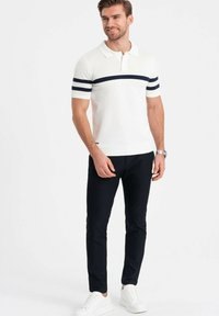 Polo shirt bianco a maglia con righe blu navy, abbinato a pantaloni scuri e sneakers bianche. Caratterizzato da un colletto classico e maniche corte.