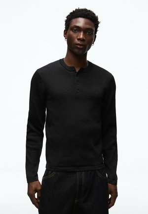 Junger Mann mit kurzen lockigen Haaren und Creolen-Ohrringen, der ein schwarzes langärmeliges Henley-Shirt und dunkle Hose vor einem weißen, einfarbigen Hintergrund trägt.
