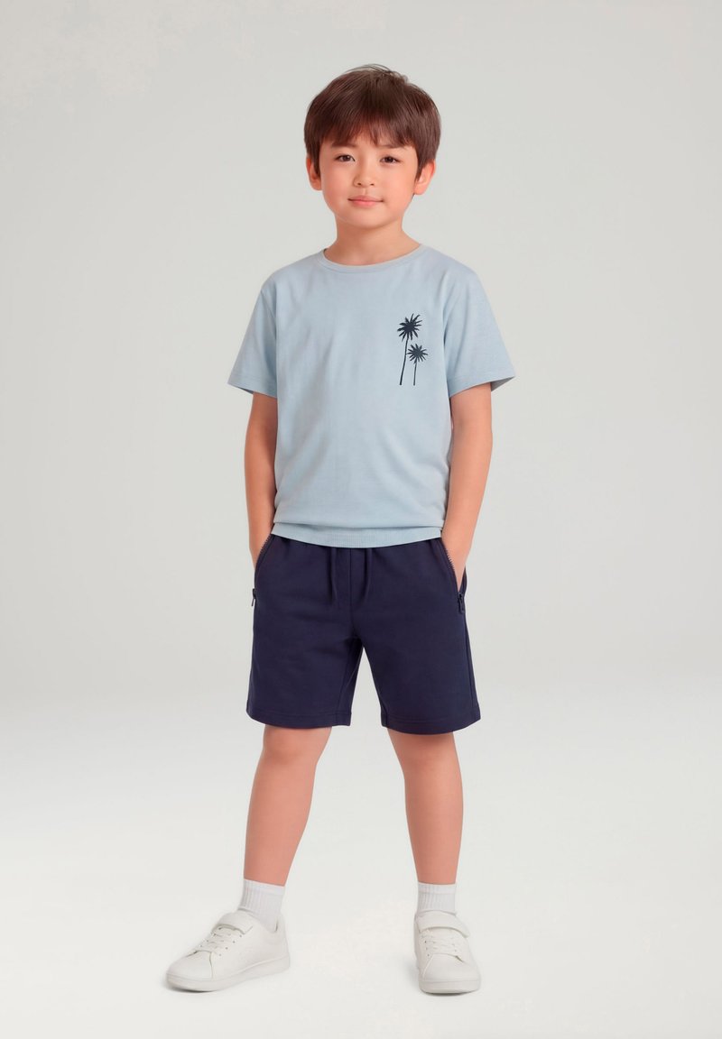 Jeune garçon debout, les mains dans les poches, portant un t-shirt bleu clair avec un imprimé de palmier, un short bleu marine, des baskets blanches et des chaussettes sur un fond clair.
