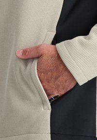 La main d’un homme reposant dans la poche d’une veste texturée beige clair avec un tissu noir visible en dessous.
