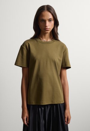 Joven mujer con el pelo largo y oscuro, lleva una camiseta de manga corta color verde oliva y una falda negra plisada sobre un fondo liso.
