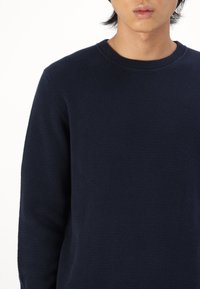 Sötétkék texturált kötött pulóver, kerek nyakkivágással és hosszú ujjakkal, diszkrét mintával, bordázott mandzsettával és alsó szegéllyel.