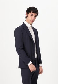 Blazer azul marino y pantalones a juego de tela suave; presenta solapas de muesca, dos botones y bolsillos laterales; llevado sobre una camisa blanca.