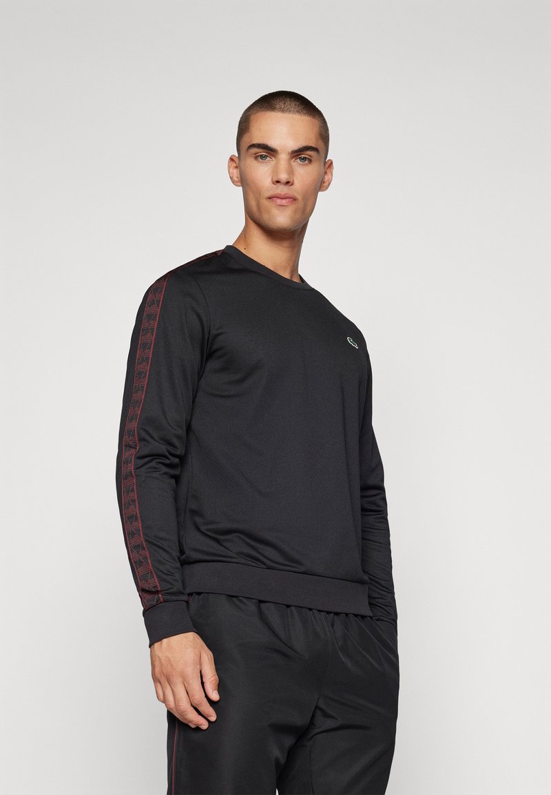 Sweatshirt noir à manches longues avec un col ras du cou classique, présentant des accents texturés rouges le long des manches et un petit logo sur la poitrine.