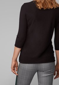 Czarna bluzka z rękawami trzyczwartowymi zestawiona z szarymi spodniami w kratę, z zielonym paskiem po bokach. Widoczna gładka faktura materiału.