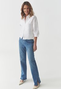 Camicia bianca con maniche tre quarti, abbinata a jeans dritti azzurri chiari con aperture laterali e tacchi a punta beige.