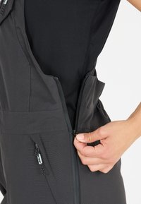 Schwarze Overall mit Reißverschluss aus strukturiertem Stoff, mit vorderem Reißverschluss und Seitentaschen. Handverstellbarer Reißverschluss auf der linken Seite.