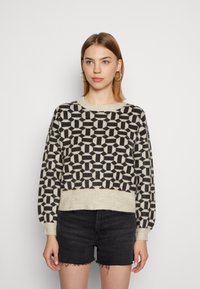 Maglione a righe nere e crema con orlo e polsini a coste, caratterizzato da design geometrici. Indossato con pantaloni corti neri e orecchini a cerchio.
