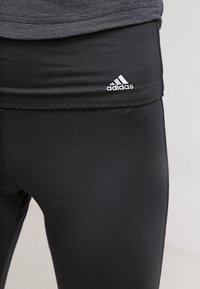 Černé sportovní legíny s přiléhavým střihem, měkkou texturou a bílým logem Adidas na pase, s podrobným šikmým stehem.