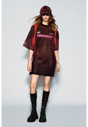 Frau trägt übergroßes dunkelrotes Tomorrowland-Hemdkleid, passende Kappe, Sonnenbrille und schwarze kniehohe Stiefel, steht vor einfarbigem Hintergrund.