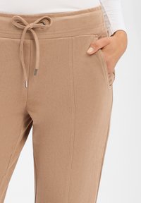 Beige jogger pants in soft fabric feature an elastic waistband with a drawstring, side pockets, and a smooth texture with no visible patterns.