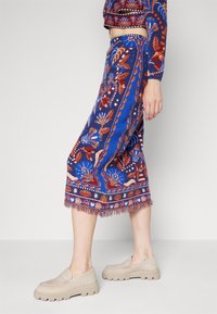 Jupe bleue imprimée à motif floral rouge, longueur genou, avec un ourlet à franges, associée à des chaussures beiges à enfiler. Le matériau semble texturé et tissé.