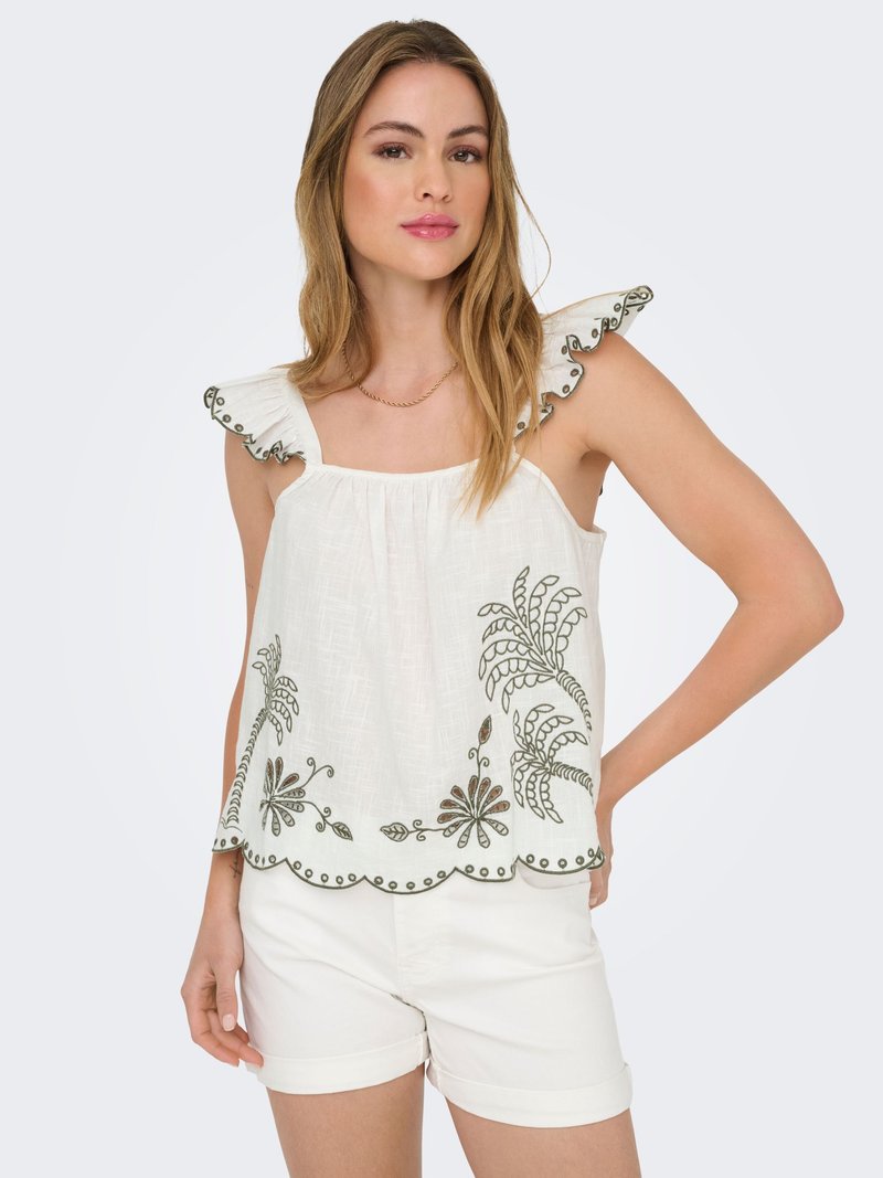 Woman wearing white sleeveless top with ruffled shoulders and floral embroidery, paired with white shorts, posing with one hand on hip.