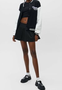 Black and white varsity jacket with logo, black fitted top, pleated black pinstripe skirt, and black chunky shoes with white socks.