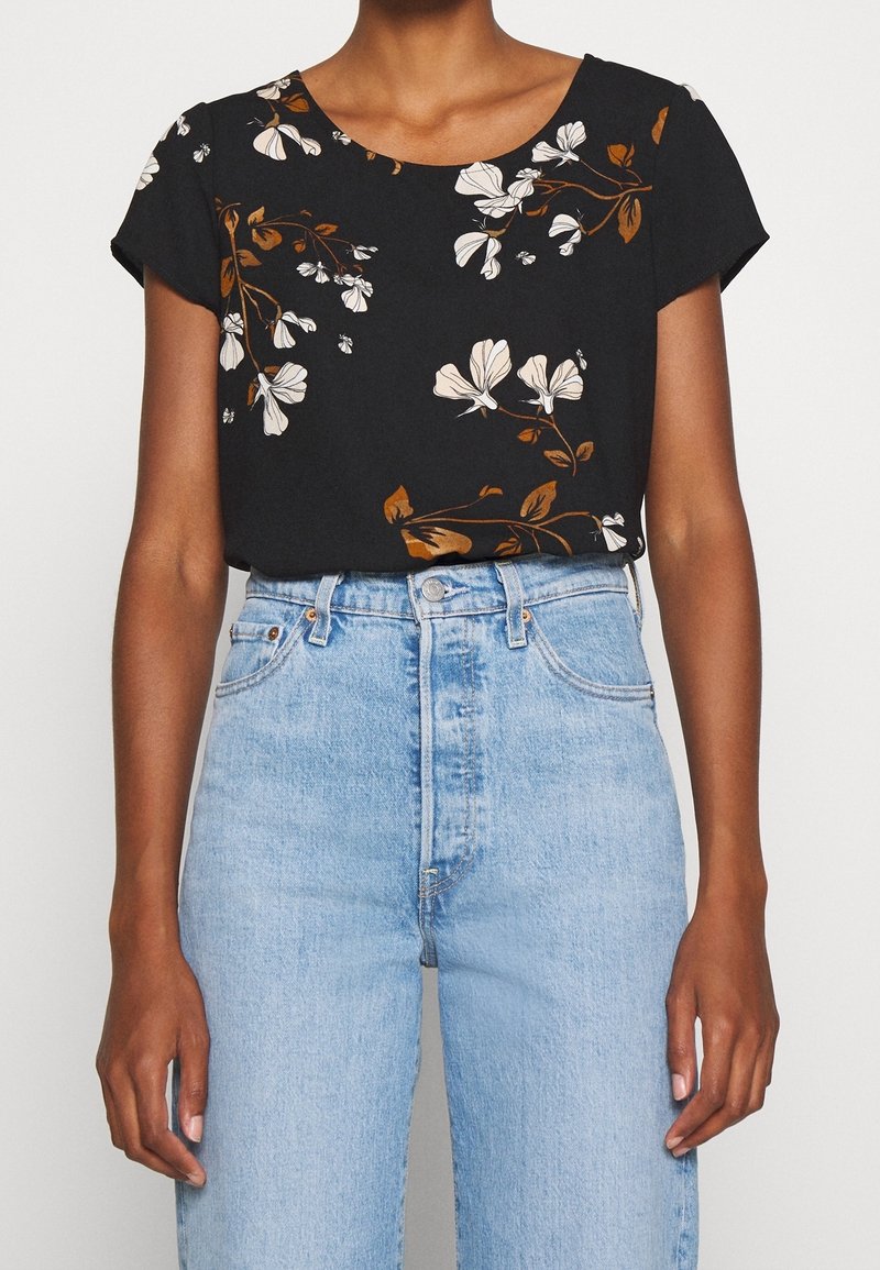 Femme portant un haut noir à manches courtes avec un imprimé floral blanc et marron rentré dans un jean taille haute bleu clair.
