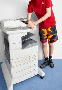Persona con una camisa roja y pantalones cortos coloridos operando una fotocopiadora de oficina blanca sobre un suelo de moqueta azul.