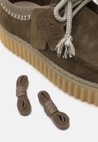 Zapato de ante marrón con un cuello de tela gris texturizada y suela de goma beige gruesa. Incluye dos pares adicionales de cordones marrones.