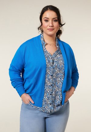 Blauwe cardigan met zakken, gebreide textuur, open voorkant, gedragen over een blauw-witte paisley-gepatroneerde blouse. Model poseert met handen in de zakken.