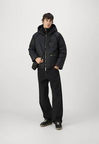 Melna polsterēta jaka ar kapuci, divi priekšējie rāvējslēdzēji, melnas džinsa bikses un melni apavi ar gaismas krāsas šņorēm; stāv pret vienkāršu fonu.