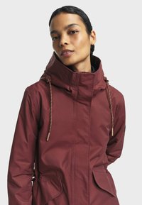 Veste à capuche bordeaux avec un col monté, des accents à cordon de serrage et deux poches avant. Fabriquée en tissu léger et résistant à l'eau.