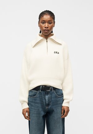Ženska s pletenicami, oblečena v bel pulover s polovičnim zadrgo z logotipom "GAP" in modre kavbojke, na enostavni svetli podlagi.