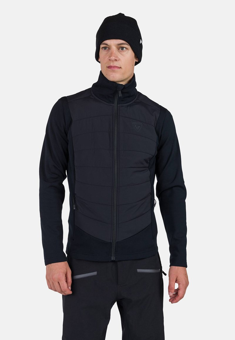 Svart isolerad väst med dragkedja fram, med en quiltad textur på torso och stretchiga ärmar. Tillsammans med en svart stickad mössa.